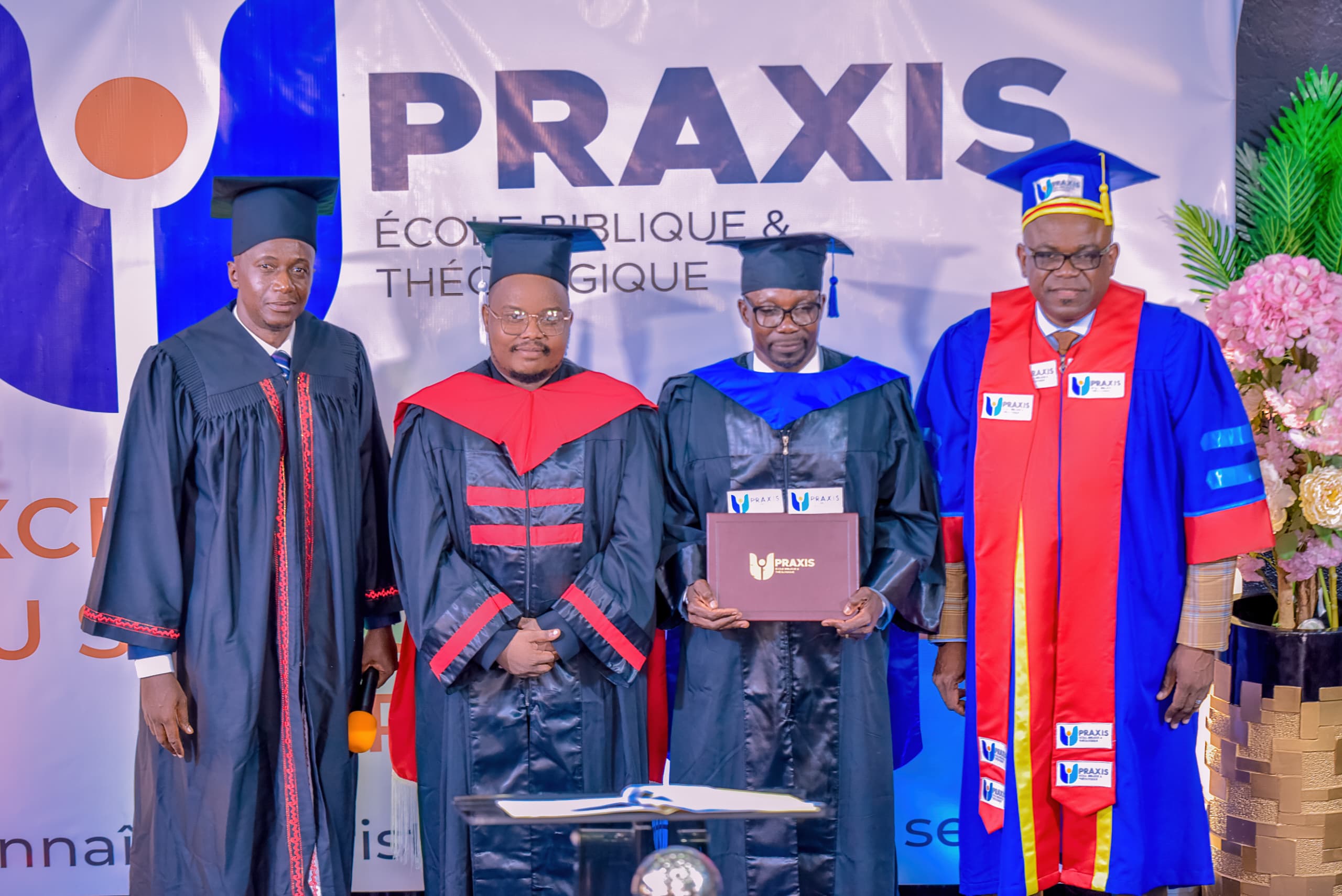 Collation des grades de la promotion Praxis : quatre diplômés en toge devant la bâche de l'École.